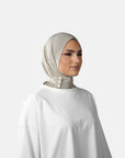Woman wearing a beige hijab and white top against a gray background